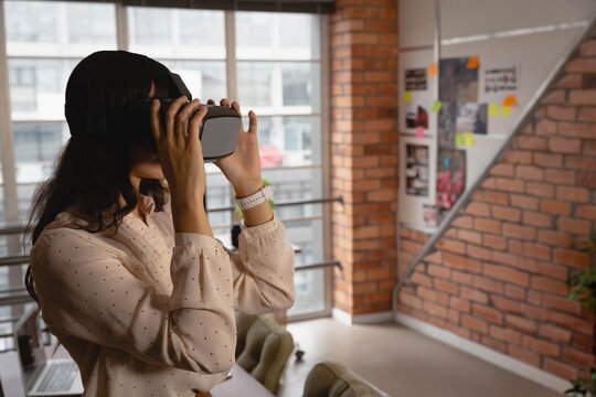 Woman holding VR headset, looking into it at office loft with smartwatch and laptop