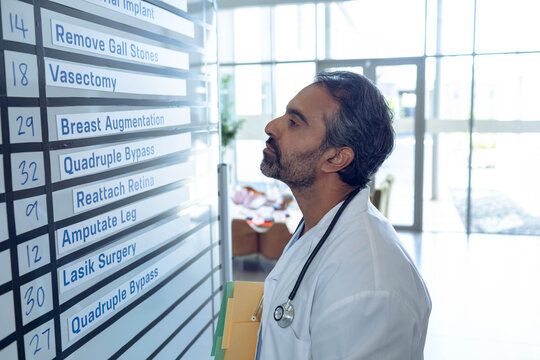 Mature Asian male doctor with stethoscope checking surgery board while holding folders by doors