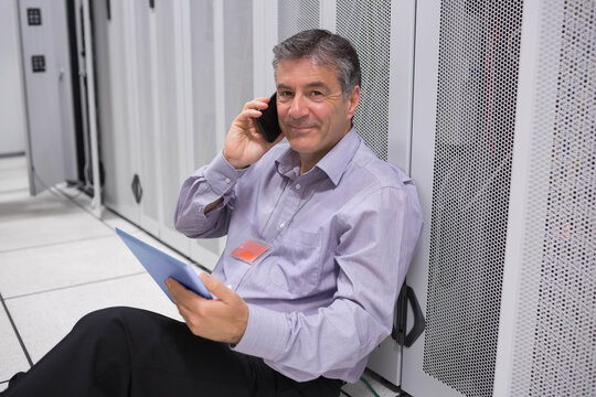 Senior man sitting on raised floor in data center, holding blue tablet and talking on smartphone
