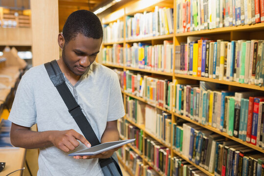 African American male tapping tablet while carrying messenger bag in library aisle, copy space
