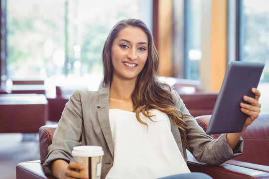 Adult woman sitting in lounge leather chair wearing grey blazer holding tablet and to-go coffee