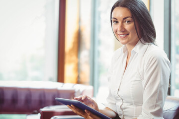Obraz premium Adult woman sitting on sofa in bright lounge wearing white blouse tapping tablet, copy space
