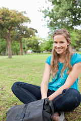 Obraz premium Woman sitting smiling on park lawn wearing turquoise blouse, jeans, gray backpack, copy space