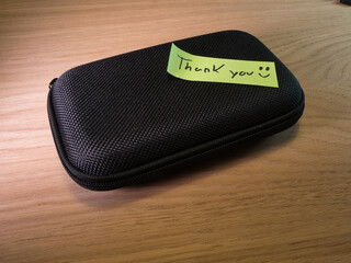Thank You Note Next to Black Hard Case - Handwritten 'Thank You' note with a smiley face next to a black zippered hard case on a light wooden desk, conveying gratitude and appreciation.