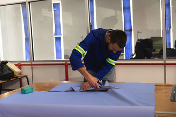 Adult man in blue jacket with neon stripes cutting light-blue fabric at textile table using shears