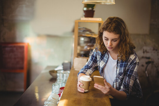 Woman sitting at wooden counter in cafe holding paper cup and checking smartphone, copy space