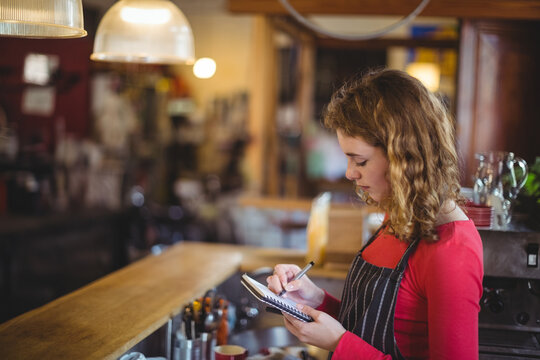 Female worker wearing striped apron writing on notepad at wooden bar, holding pen left, copy space