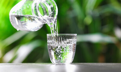 Pouring water into glass from jug at grey table against blurred green background, closeup