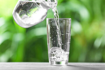 Pouring water into glass from jug at grey table against blurred green background, closeup