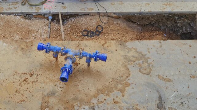 Top view of a muddy trench in a road construction site. A blue ductile iron water pipe T-joint is visible sitting in the mud and water, ready for installation. Construction tools, including a jackhamm
