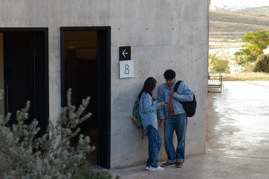 Young students standing by a concrete wall on campus during daylight, holding smartphones, staying connected and interacting with technology for education or social media
