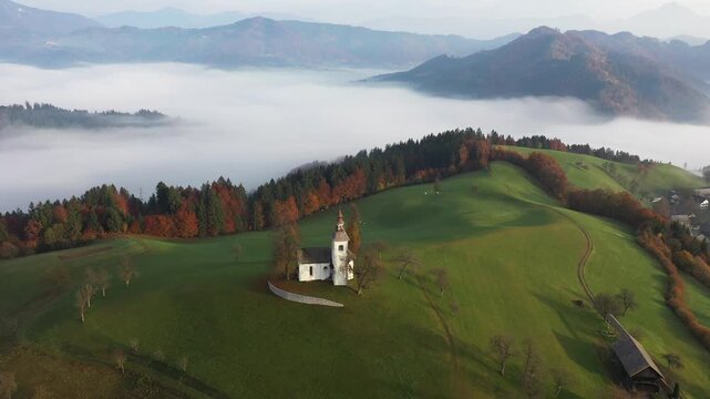 Aerial view of a solitary white church perched atop a vibrant green hill, framed by a forest ablaze with autumn colors and a distant sea of fog, Sv.Tomaz, Ormoz, Slovenia.