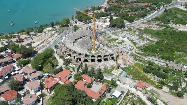Aerial view of the ancient Side Antique Theatre featuring stone ruins and construction cranes amid the blend of turquoise sea and lush greenery, Side, Antalya, Turkey.