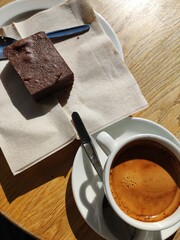 Espresso and Brownie on Wooden Table. A cup of rich espresso sits beside a chocolate brownie on a wooden table, both catching the warm sunlight. The scene suggests a relaxing coffee break.