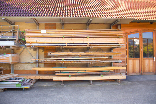 Stacked lumber stored outside a wooden workshop building with large glass doors