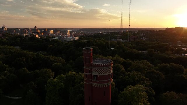 Aerial view of red brick water tower in Eden Park, contrasting with the lush greenery and distant city skyline at sunset, Cincinnati, Ohio, United States.