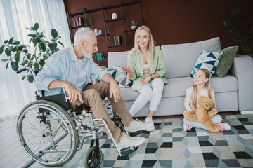 Heartwarming family moment at home with grandfather in a wheelchair talking with mom and daughter on a cozy sofa in a bright living room while plants