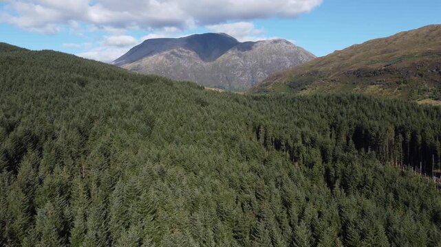 Aerial view of lush green forestation sprawling across hillsides, contrasting with a distant, rugged mountain range under a partly cloudy sky, Fort William, Scotland, United Kingdom.