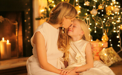Happy mother kissing her child daughter near Christmas tree and fireplace at home
