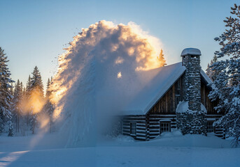 Snow blows off the roof of a log house with a sunlight effect