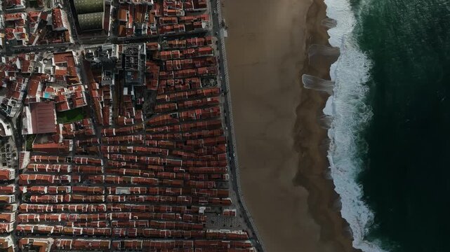Aerial view of the city buildings with red tile roofs meeting the sandy beach and the dark blue ocean with white waves, Nazare, Leiria District, Portugal.