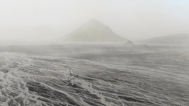 Aerial view of a lone car traversing the braided river system against the backdrop of a mountain shrouded in mist, Skaftarhreppur, Iceland.