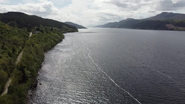 Aerial view of dark, rippling waters of Loch Ness, contrasting with the lush green forests lining its shores under a cloudy sky, Inverness, Scotland, United Kingdom.