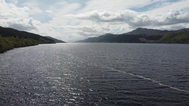 Aerial view of the dark, rippling waters of Loch Ness, embraced by verdant hills under a sky dappled with fluffy clouds, Inverness, Scotland, United Kingdom.