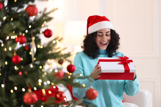 Happy woman with Santa hat and gift near Christmas tree at home - Powered by Adobe