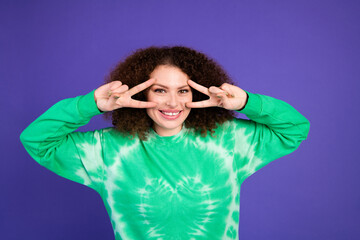 Young woman in a green tie dye sweater smiles and makes a peace sign against a purple backdrop for a stylish lifestyle stock photo