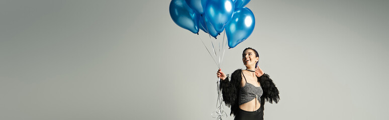 Beautiful young woman in stylish attire joyfully holding blue balloons in a minimalist setting