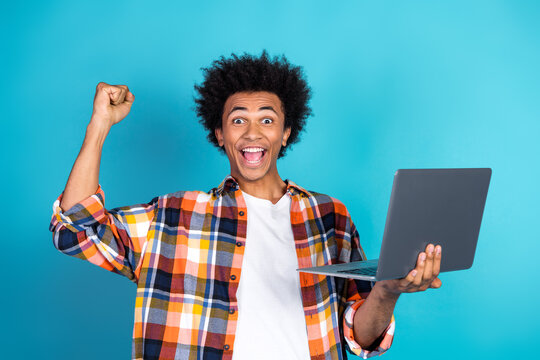 Young man with curly hair celebrates success holding laptop in a bright blue studio with a fist pump and big smile