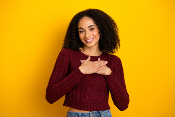 Smiling young woman in a burgundy sweater stands against a bright yellow background with hands over heart conveying joy and friendly vibe