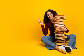 Young mixed race woman glasses sits on yellow background holding tall stack of book points with a cheerful smile to show study life learning creativity