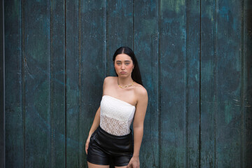 Fototapeta premium Young woman, attractive, brunette, blue eyes, Hispanic, wearing a white top and leather shorts, posing and looking at the camera leaning against a blue door in the background. Concept: model, beauty.