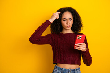 Young mixed race woman against yellow background wearing a maroon sweater and checks her smartphone with puzzled expression while shopping