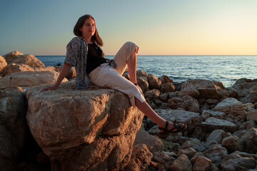 A girl poses and relaxes by the sea, sitting on rocks near the slope of a high cliff, panoramic view of the seashore and mountains at sunset
