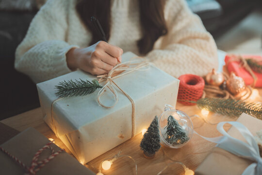 Woman writing a greeting on a Christmas gift box decorated with natural twine and pine branch, capturing festive preparation, holiday crafting, warm seasonal gifting and joyful celebration moments - Powered by Adobe