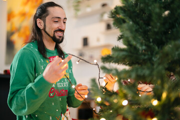 A man in a festive sweater decorates a Christmas tree with lights, smiling as he works.