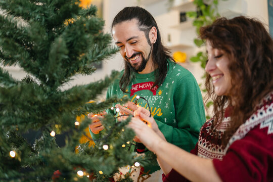 A smiling couple decorates a Christmas tree with lights, sharing a festive moment together.