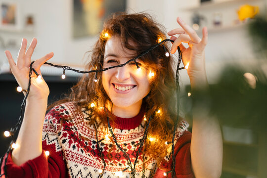 A young woman with curly hair smiles as she holds decorative lights around her face and hair, creating a festive and joyful atmosphere.