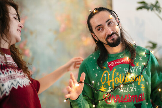 A man and woman share a festive moment, entangled in twinkling holiday lights. The man wears a green sweater with a holiday message, while the woman's hand reaches out.