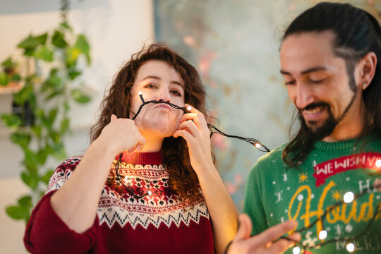A woman playfully uses string lights as a mustache while a man in a festive sweater smiles.