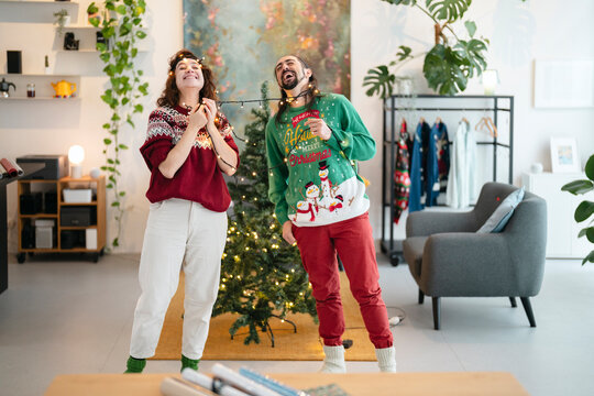 A joyful couple in festive sweaters laugh while tangled in Christmas lights next to a decorated tree, celebrating the holiday season together. - Powered by Adobe