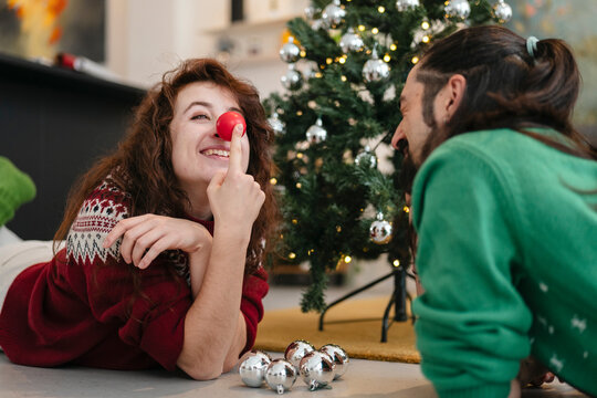 A woman with a red clown nose playfully touches her nose while looking at a man near a decorated Christmas tree.