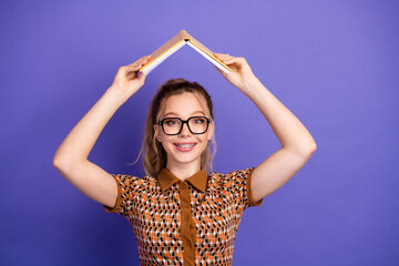 Young student girl with glasses holds a book over her head like a roof smiles against a violet...
