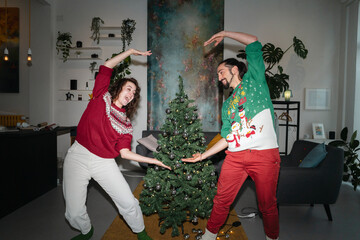 A joyful couple in festive sweaters dances around a decorated Christmas tree, celebrating the holiday season together in a dimly lit room.