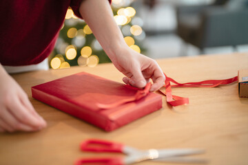 A person's hands tie a red ribbon around a gift box on a wooden table, with a blurred Christmas tree in the background.