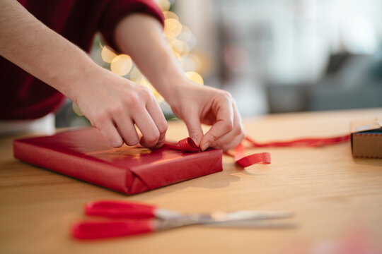 Hands carefully wrap a red gift box with ribbon, with scissors nearby and festive lights in the background.