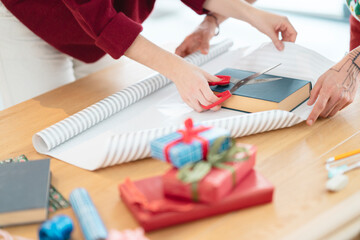 Two people are wrapping gifts on a wooden table, one person is cutting wrapping paper with scissors, the other is holding it.